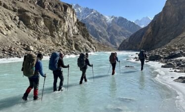 Trekkers crossing a wide shallow glacial river on the Shimshal Pass trek, turquoise water and dramatic rocky mountain valley, northern Pakistan