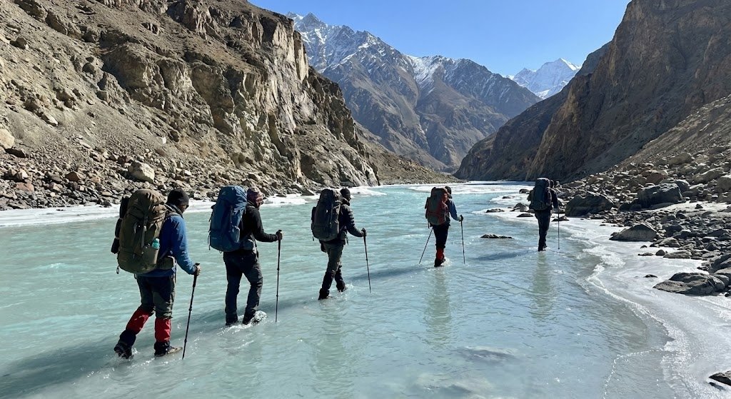 Trekkers crossing a wide shallow glacial river on the Shimshal Pass trek, turquoise water and dramatic rocky mountain valley, northern Pakistan