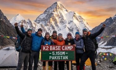 A group of trekkers celebrating at K2 Base Camp, posing behind a red and green wooden sign that reads "K2 BASE CAMP - 5,150M - PAKISTAN" with the mountain peak glowing in the sunset behind them