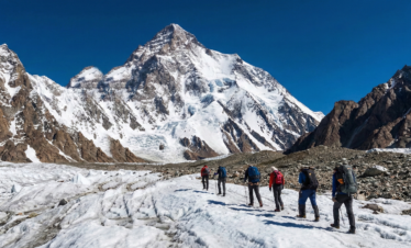 A group of trekkers walking on the white, cracked surface of a glacier, heading directly toward the massive, snow-covered face of K2 under a brilliant blue sky.