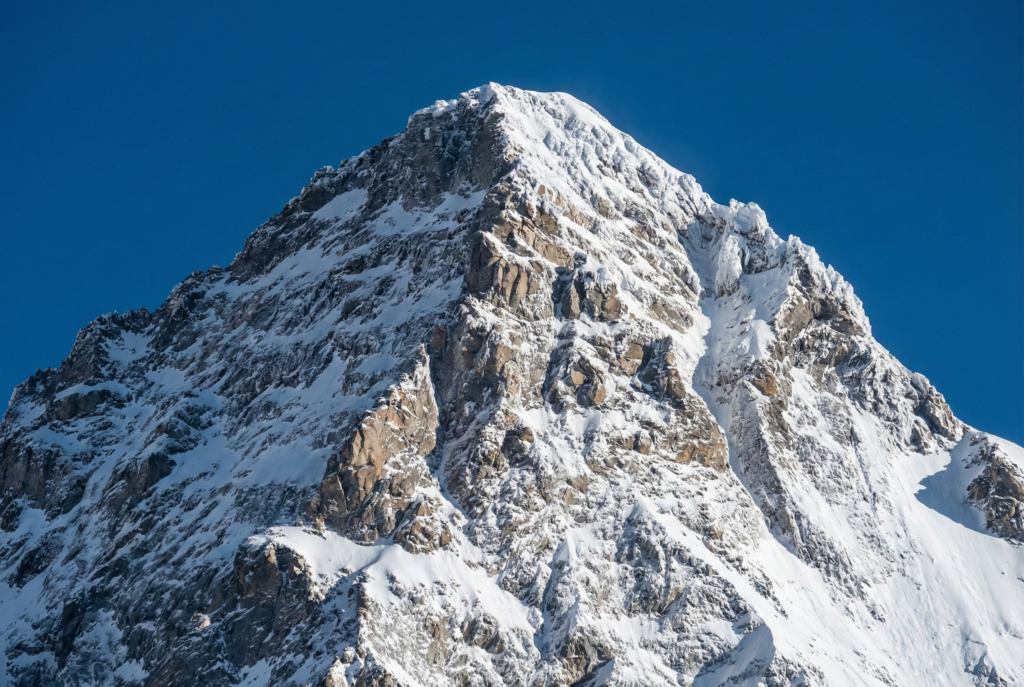 A high-resolution close-up of the K2 summit, showcasing the sharp, icy ridgelines and steep rocky faces of the "Savage Mountain" against a piercing, cloudless blue sky.