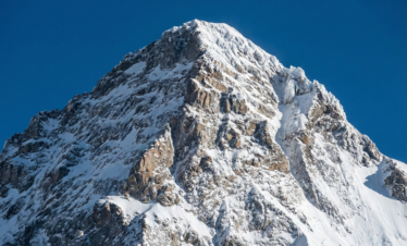 A high-resolution close-up of the K2 summit, showcasing the sharp, icy ridgelines and steep rocky faces of the "Savage Mountain" against a piercing, cloudless blue sky.