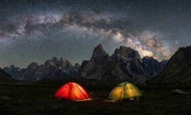 Expedition tents glowing under a dramatic Milky Way sky above the granite peaks of Nangma Valley, high altitude trekking Skardu Pakistan