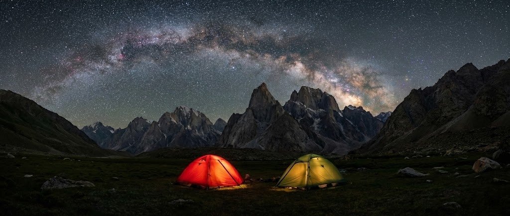 Expedition tents glowing under a dramatic Milky Way sky above the granite peaks of Nangma Valley, high altitude trekking Skardu Pakistan