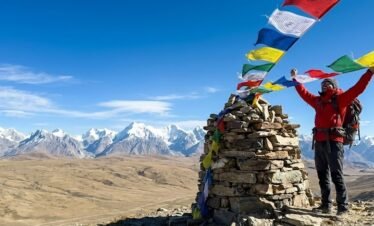 Trekker standing at the summit cairn of Shimshal Pass at 4,735m with prayer flags and vast Pamir plateau stretching behind, northern Pakistan