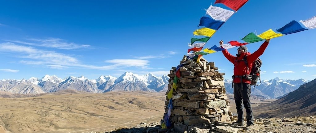 Trekker standing at the summit cairn of Shimshal Pass at 4,735m with prayer flags and vast Pamir plateau stretching behind, northern Pakistan
