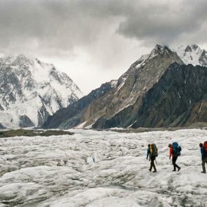 A group of four trekkers with large backpacks walking across the vast, rugged white ice of the Baltoro Glacier under a dramatic, overcast sky with jagged rocky peaks in the background.