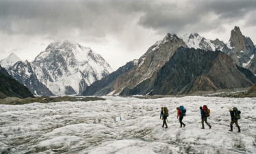 A group of four trekkers with large backpacks walking across the vast, rugged white ice of the Baltoro Glacier under a dramatic, overcast sky with jagged rocky peaks in the background.