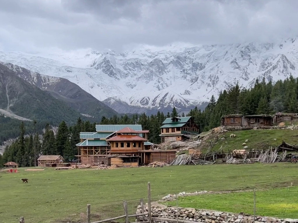 A traditional mountain lodge overlooks the sprawling meadows of Fairy Meadows in Gilgit-Baltistan, Pakistan, with snow-capped peaks of the Nanga Parbat massif rising dramatically in the background. This high-altitude guesthouse serves as the base camp and accommodation hub for trekkers pursuing the Nanga Parbat Base Camp trek and exploring the alpine meadows of northern Pakistan. The lodge provides warm hospitality at over 3,600 metres elevation amid coniferous forests and mountain vistas.