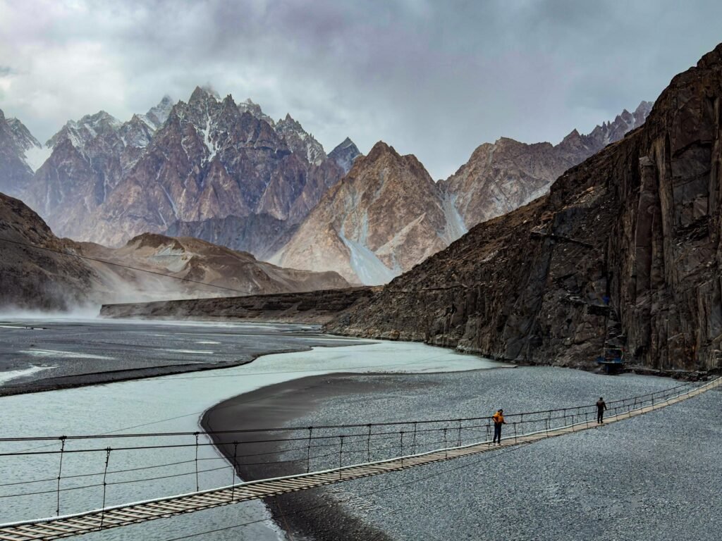 Two trekkers crossing the Hussaini Hanging Bridge over the glacial Hunza River with the Passu Cones mountain spires rising behind them in Upper Hunza, Gilgit-Baltistan, Pakistan